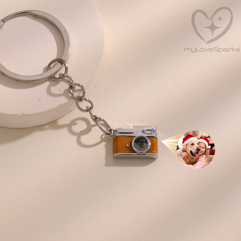 May include: A keychain featuring a miniature camera charm in orange and silver. The charm is attached to a chain and a keyring. A small photo is projected from the camera. The logo "myLoveSparks" is in the upper right corner.