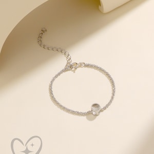 K&ouml;nnte beinhalten: Ein zartes Silberarmband mit einem kleinen, klaren, runden Anh&auml;nger. Das Armband besteht aus kleinen, funkelnden Perlen und hat eine verstellbare Kette. Der Hintergrund ist hellbeige.