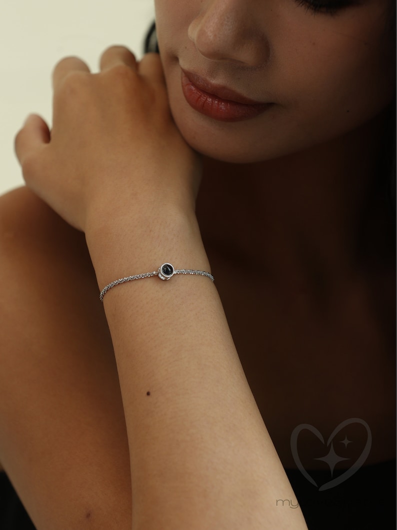 K&ouml;nnte beinhalten: Ein filigranes Silberarmband mit einem kleinen, runden, schwarzen Edelstein in der Mitte. Das Armband hat ein schlichtes Kettendesign, und der Edelstein scheint in einer L&uuml;nette gefasst zu sein. Das Armband wird am Handgelenk getragen.