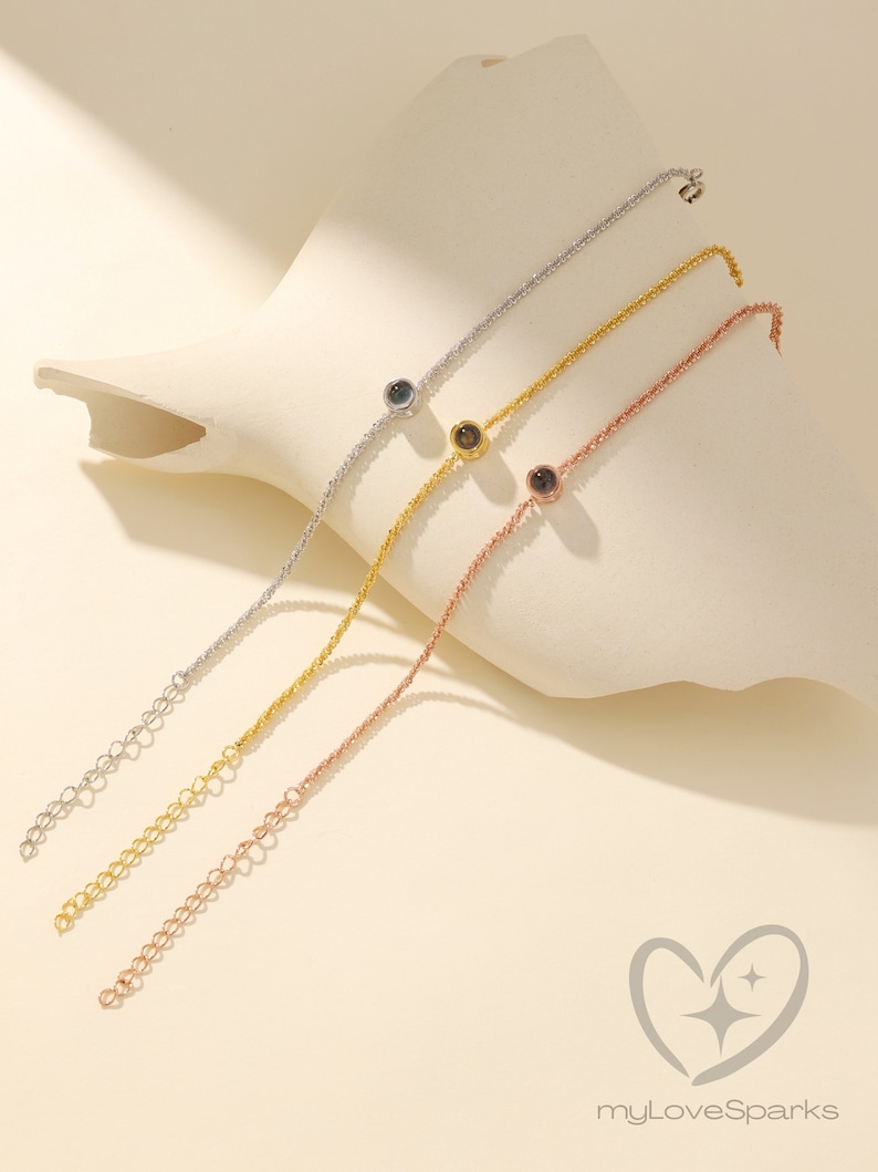 K&ouml;nnte beinhalten: Drei zarte Kettenarmb&auml;nder in Silber, Gold und Ros&eacute;gold, jedes mit einem kleinen, runden Anh&auml;nger. Die Armb&auml;nder werden auf einer wei&szlig;en, abstrakten Form vor einem weichen, neutralen Hintergrund pr&auml;sentiert. Die Armb&auml;nder sind in der L&auml;nge verstellbar.