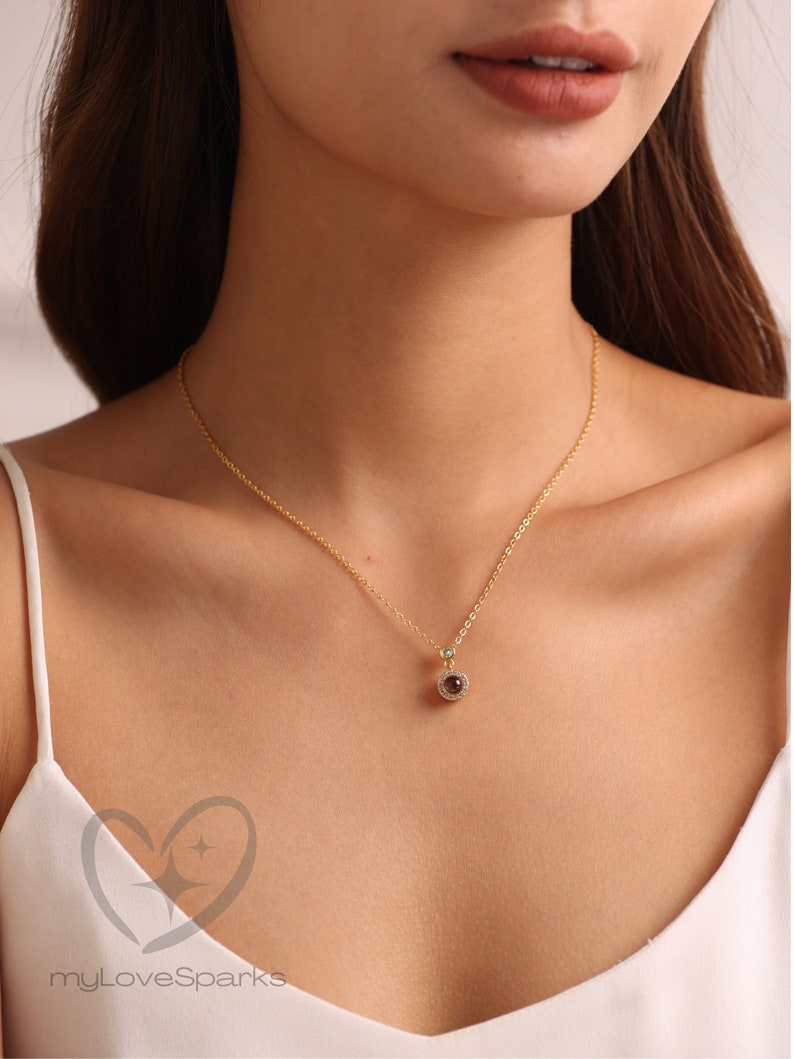 May include: A gold necklace with a circular pendant. The pendant has a dark central stone encircled by smaller, clear stones. The necklace is worn by a person wearing a white top. The necklace is a delicate piece.