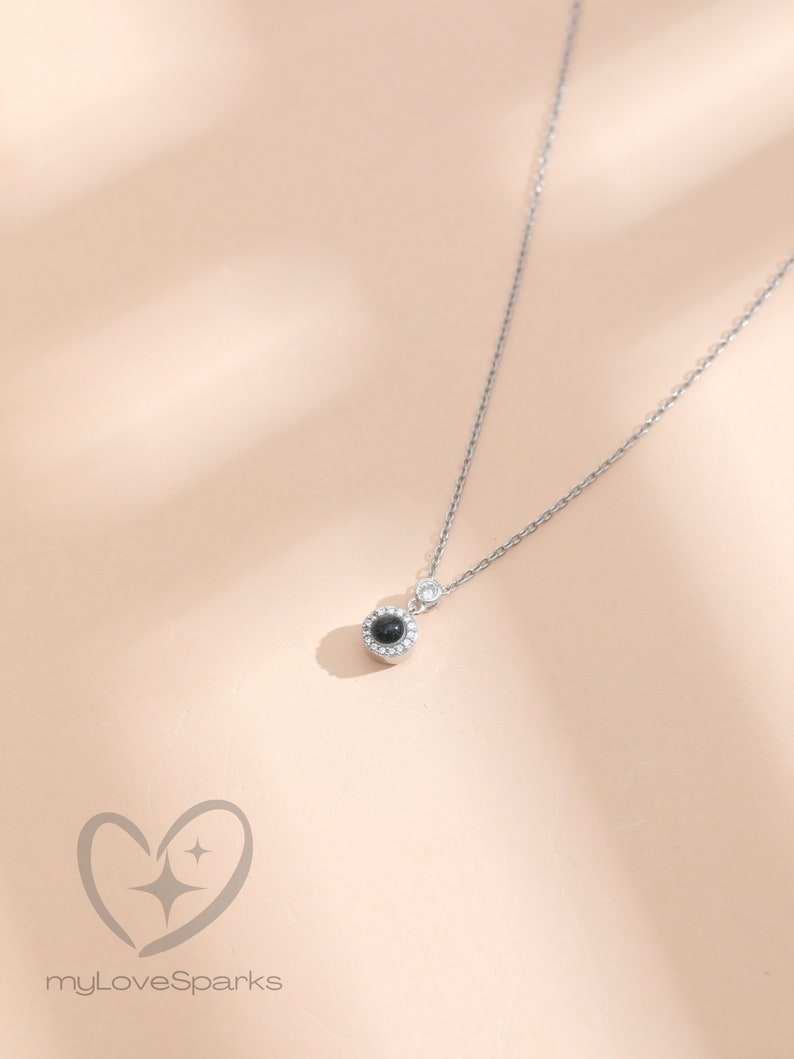 May include: A silver necklace featuring a small, round, black pendant surrounded by small clear stones. The delicate chain has a small, clear stone near the clasp. The logo "myLoveSparks" is at the bottom.