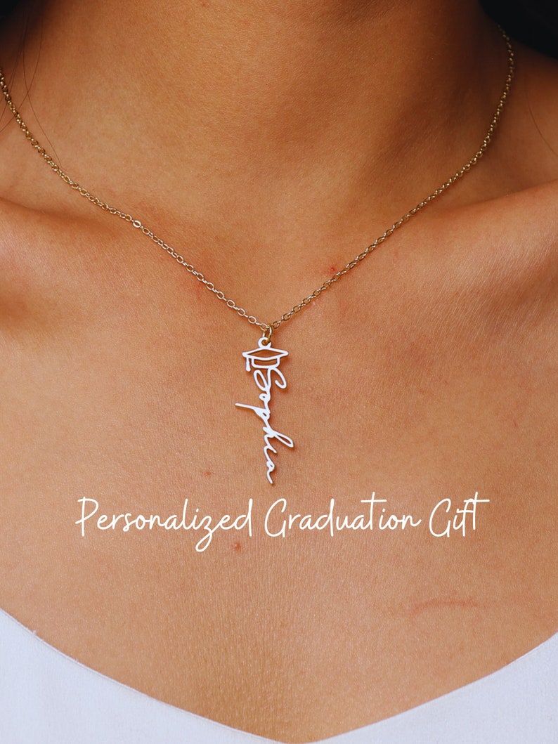 May include: A gold chain necklace with a pendant featuring a graduation cap and the name "Sophia" written in cursive. The necklace is worn by a person with a white shirt. The text "Personalized Graduation Gift" is written below the necklace.