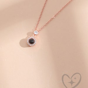 May include: Rose gold necklace with a pendant featuring a dark stone surrounded by small clear stones. A single clear stone is set higher on the chain. The necklace is displayed on a light peach background.