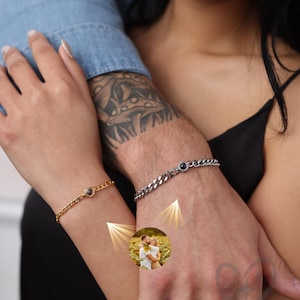 May include: A couple wearing matching bracelets with a small photo inside the charm. The bracelets are silver and gold with a black stone in the center of the charm. The photo shows a couple kissing in a field of flowers. The text "myLoveSparks" is visible in the bottom right corner of the image.