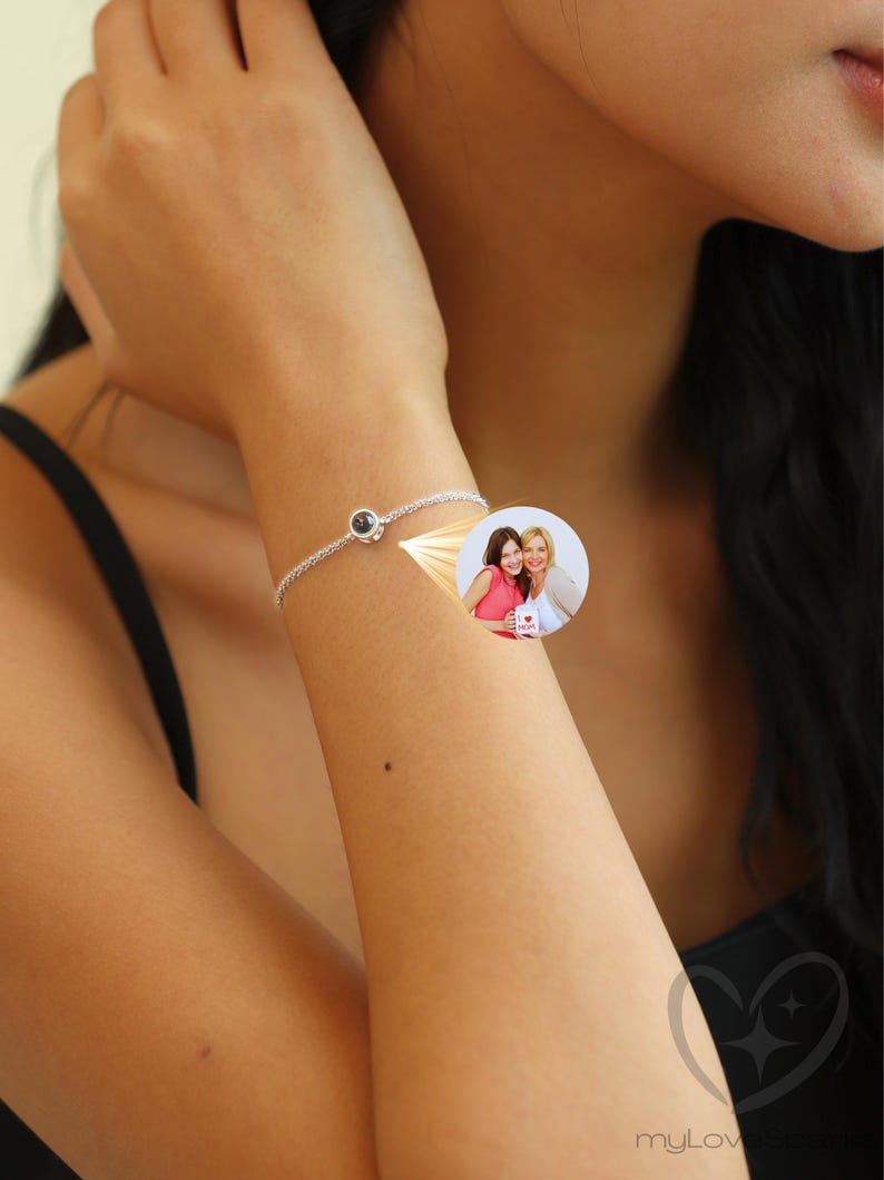 K&ouml;nnte beinhalten: Ein silbernes Armband mit einem kleinen, runden, dunkel gef&auml;rbten Stein. Das Armband befindet sich an einem Handgelenk, mit einem kleinen Bild von zwei Frauen im Hintergrund. Das Armband ist filigran und aus Metall.