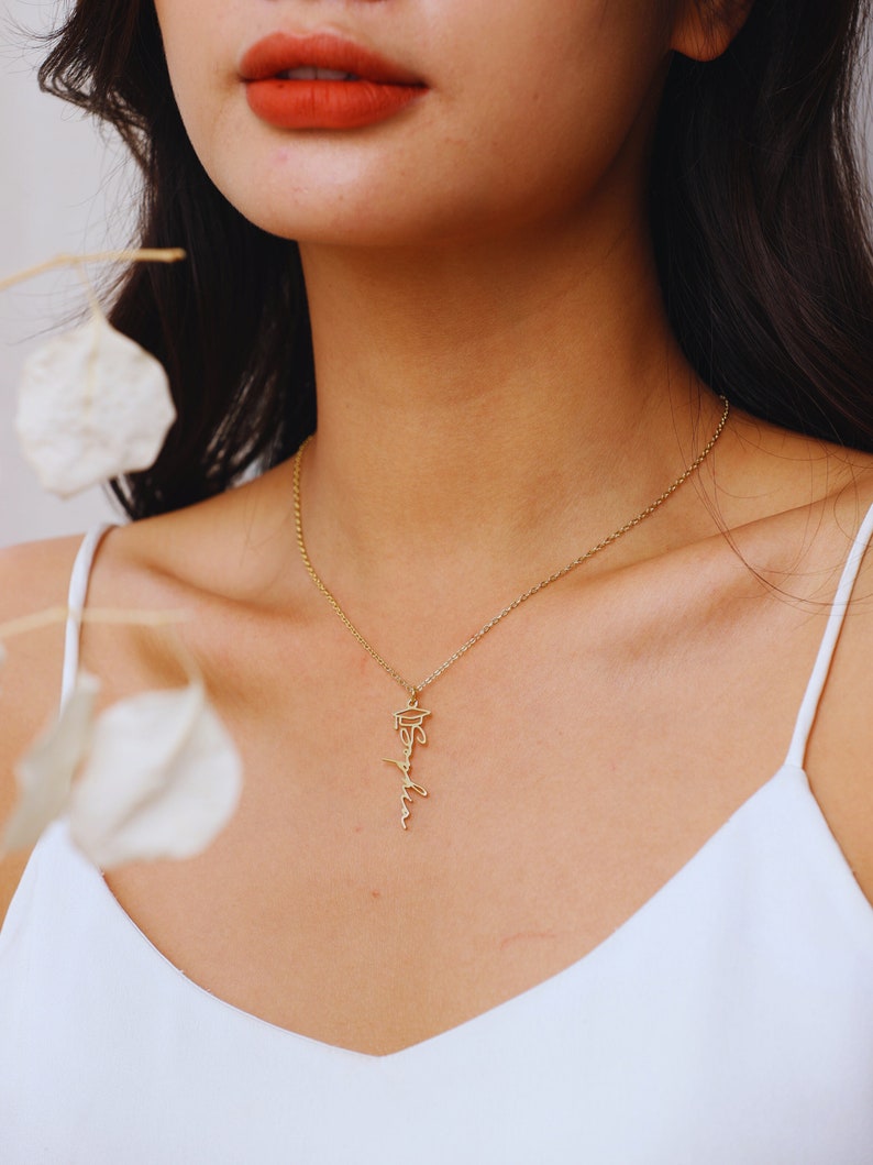 May include: A gold chain necklace with a delicate pendant featuring the word "Graduate" in cursive script. The pendant is a graduation cap.