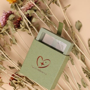 May include: A green gift box with a heart-shaped logo that says "myLoveSparks" and a clear window on the top. The box is surrounded by dried flowers.