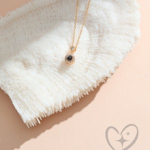 May include: A gold necklace with a circular pendant containing a black stone and clear crystals. A single clear crystal sits above the pendant. The necklace is presented on a white, textured surface, with a gold chain.