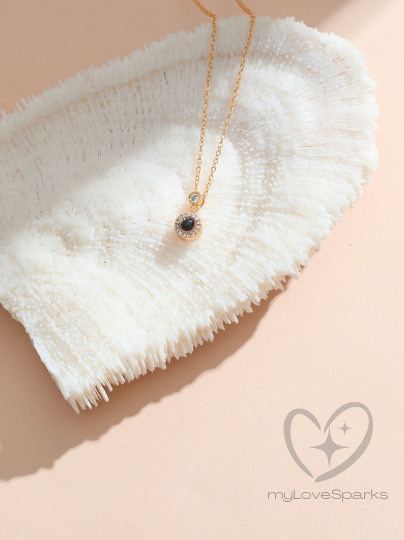May include: A gold necklace with a circular pendant containing a black stone and clear crystals. A single clear crystal sits above the pendant. The necklace is presented on a white, textured surface, with a gold chain.