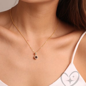 May include: A gold necklace with a small, round pendant. The pendant has a dark red gemstone surrounded by a halo of smaller, clear stones. The necklace is worn against a white top.