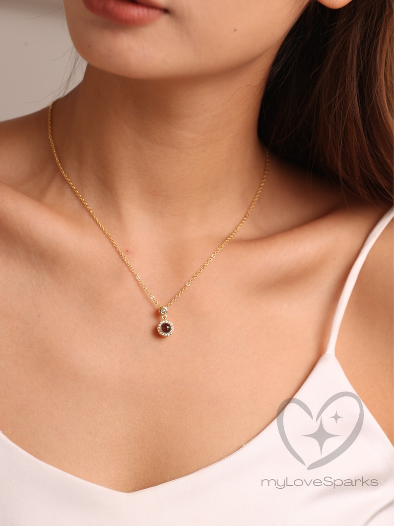 May include: A gold necklace with a small, round pendant. The pendant has a dark red gemstone surrounded by a halo of smaller, clear stones. The necklace is worn against a white top.