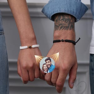 May include: Two people holding hands, each wearing a black and white string bracelet with a small bead. A photo of a couple is superimposed on the man's wrist, with a golden glow effect.