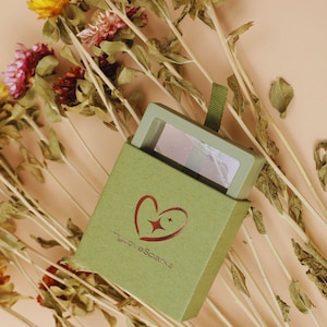 May include: A small, open green box with the text "myLoveSparks" and a heart logo, revealing a small makeup compact. The box is surrounded by dried flowers with yellow, pink, and brown hues, set against a peach background.