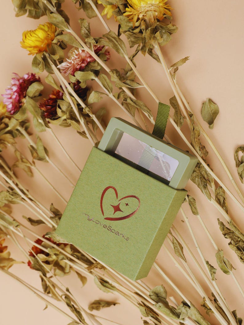 May include: A small, open green box with the text "myLoveSparks" and a heart logo, revealing a small makeup compact. The box is surrounded by dried flowers with yellow, pink, and brown hues, set against a peach background.