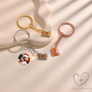 May include: Three keychains with miniature camera charms. One has a gold ring and chain, another a silver ring and chain, and the third a rose gold ring and chain. The camera charms are orange and silver. One keychain has a photo of two people.