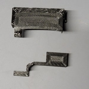 May include: Two black 3D printed plastic pieces. The larger piece is rectangular with a grid pattern. The smaller piece is a curved shape with a rectangular section.