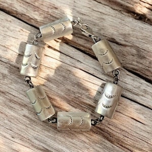 May include: A silver bracelet with a chain and cylindrical beads. Each bead has a repeating pattern of half-circles.
