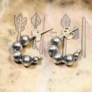 May include: A pair of silver-colored hoop earrings. Each earring is composed of a series of graduated spheres, creating a curved shape. The earrings have a post and butterfly backing. The background is a textured, tan surface with etched arrow designs.
