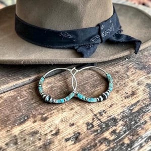 Puede incluir: Pendientes de aro plateados adornados con cuentas turquesas y plateadas. Los pendientes se exhiben sobre una superficie de madera rústica, con un sombrero marrón y una banda negra en el fondo. Estilo bohemio.