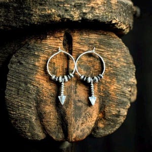 May include: Silver hoop earrings with a beaded design and arrow pendants. The earrings are displayed on a textured, dark brown wooden surface. The earrings have a bohemian or tribal aesthetic.