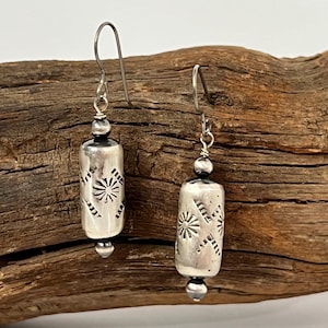 May include: A pair of silver earrings with a geometric design. The earrings are made of sterling silver and have a cylindrical shape. The earrings are hanging from a wooden surface.