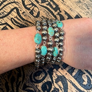 May include: A stack of silver-coloured beaded bracelets with turquoise oval stones. The bracelets are worn on a wrist, showcasing the contrast between the silver beads and the bright blue-green stones. The bracelets are on a wooden surface.
