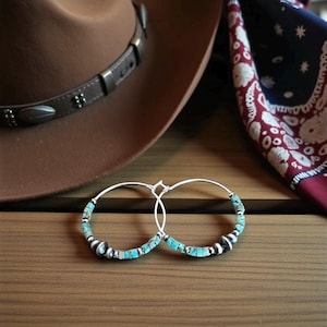 May include: A pair of silver hoop earrings featuring small, rectangular turquoise stone beads and round silver accent beads. The earrings are positioned in front of a brown hat with a leather band and a red, white, and blue patterned bandana.