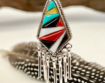 May include: A silver pendant with a geometric design featuring turquoise, red, black, white, and yellow stones. The pendant has a fringe of silver beads.