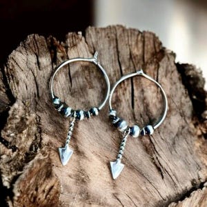 May include: A pair of silver hoop earrings with a small arrow charm dangling from each hoop. The hoops are adorned with a row of small silver beads.