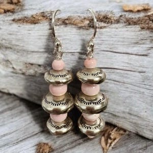 Könnte beinhalten: Ohrringe zum Hängen mit silberfarbenen, gemusterten Perlen und weichen, rosa, runden Perlen. Die Ohrringe hängen an silbernen Ohrhaken und erzeugen einen geschichteten, böhmischen Stil. Ein einzigartiges Accessoire.