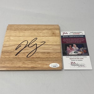 Puede incluir: Una placa de madera con una firma oscura y un certificado de autenticación JSA. La placa tiene un patrón de vetas de madera de color marrón claro. El certificado incluye el texto "BASIC CERTIFICATION" y "JSA Authentication".