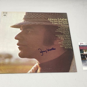 May include: A vintage vinyl record album cover featuring a black and white photo of Johnny Mathis. The album title is "The First Time Ever (I Saw Your Face)" and includes a list of songs on the back cover. The album is signed by Johnny Mathis.