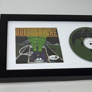 May include: Framed CD and album art featuring the band Queensrÿche. The album art has a green and brown color scheme with the band's name. The CD is green with a black bat logo. The frame is black.