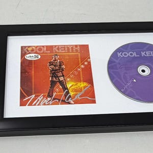 以下が含まれることがあります： Kool KeithのCDとアルバムアートを額装した音楽記念品。アルバムアートは、赤とオレンジの背景に男性が描かれています。CDは紫で、アーティスト名が書かれています。黒いフレームがアイテムを囲んでいます。