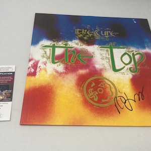 May include: A signed The Cure album titled "The Top" with a colorful abstract background and a gold seal. The signature is in black ink.