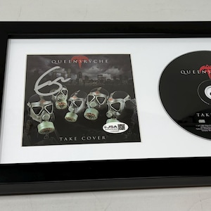 May include: Framed Queenrÿche "Take Cover" CD and album art. The album art features gas masks and an autograph. The CD is black with red text. A JSA Basic Certification sticker is in the lower left corner.