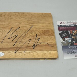 May include: A wooden basketball court floorboard signed in black marker by a basketball player. The floorboard is a light brown colour and has a grain pattern. The signature is clear and legible. The floorboard is accompanied by a JSA authentication card with the certification number VV42103.