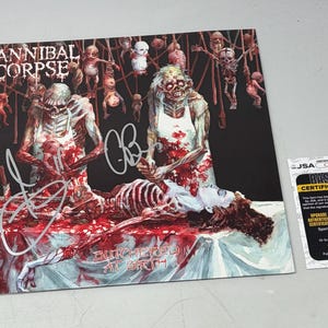 Puede incluir: Una impresión de la portada del álbum Butchered at Birth de Cannibal Corpse con una ilustración gráfica de dos figuras con delantales blancos sobre un esqueleto, figuras colgantes al fondo y una firma plateada, junto a una tarjeta de certificación JSA.