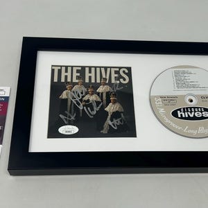 May include: Framed music memorabilia featuring a CD and a signed photo of The Hives. The CD is silver with black text, and the photo is black and white. A JSA authentication certificate is included.
