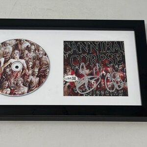 Puede incluir: Un marco negro que contiene un CD de Cannibal Corpse The Bleeding y una portada de álbum autografiada. El conjunto incluye un paspartú blanco con el disco a la izquierda y la portada firmada a la derecha. Se incluye una tarjeta de certificación JSA.