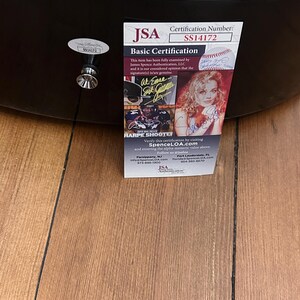 May include: An acoustic guitar with a JSA Basic Certification label. The label displays the certification number SS14172, a signature, and the text "HARPE SHOOTER". The guitar rests on a wooden surface.