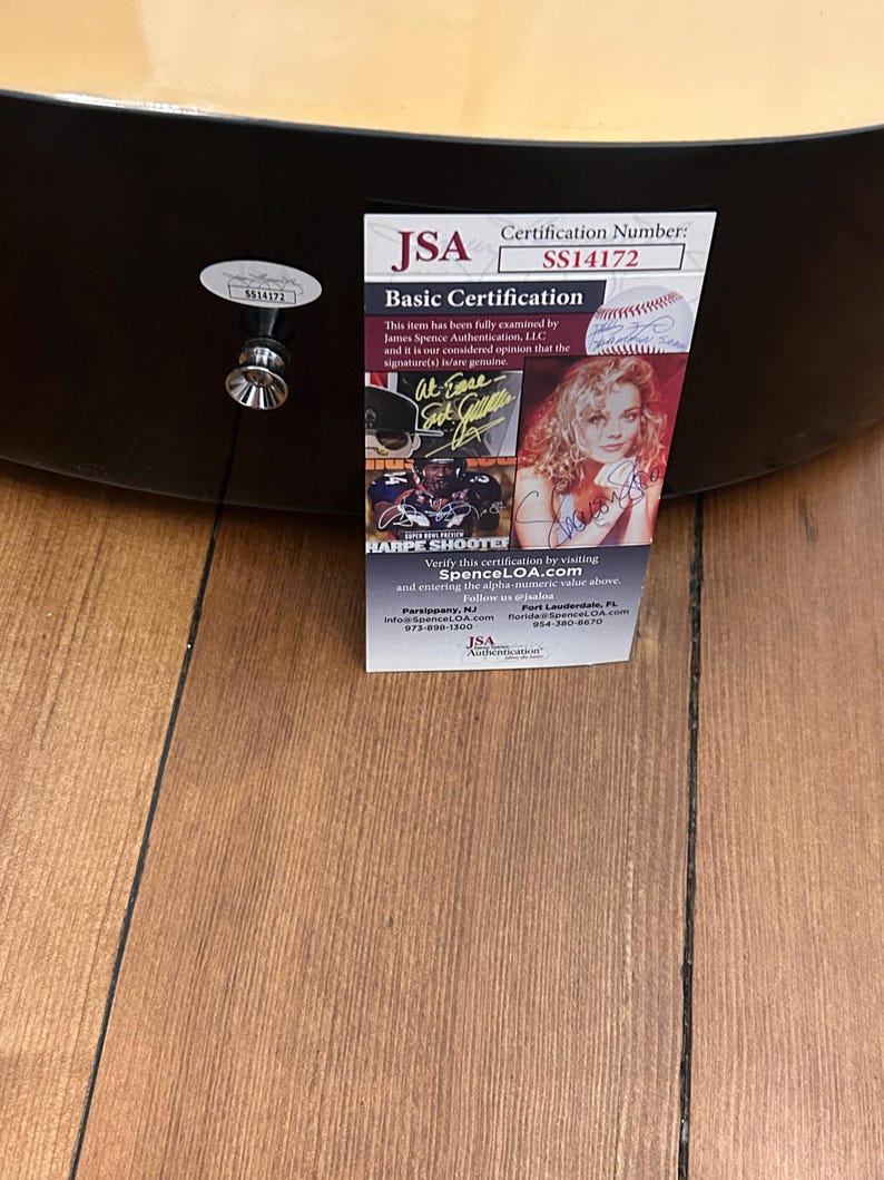 May include: An acoustic guitar with a JSA Basic Certification label. The label displays the certification number SS14172, a signature, and the text "HARPE SHOOTER". The guitar rests on a wooden surface.