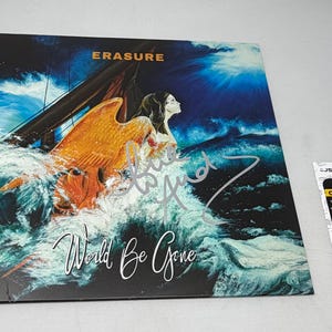 Może przedstawiać: Okładka albumu "World Be Gone" zespołu Erasure przedstawiająca kobietę z pomarańczowymi skrzydłami pośród burzliwych fal. Grafika jest podpisana i zawiera tekst "World Be Gone". Widoczna jest również naklejka certyfikacyjna JSA.