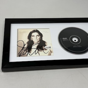 Puede incluir: CD enmarcado y portada del álbum con el retrato y la firma de una mujer. El CD es negro y el marco es negro con un passe-partout blanco. Una pegatina de certificación está a la izquierda.