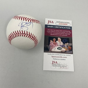 Puede incluir: Una pelota de béisbol blanca con costuras rojas y una firma azul. Una tarjeta de certificación básica de JSA con el número de certificación AM10606 está al lado de la pelota.