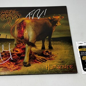May include: Album cover for Cattle Decapitation's "Humanure." The artwork depicts a brown cow with a gruesome wound, standing on a field of gore. The album title is in red. The cover is signed in white. A certification sticker is on the right.