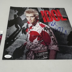 May include: A signed photograph of Billy Idol, a rock musician, wearing a white and red patterned shirt with a black belt. The background is a dark gray with a blurry image of a person in the background. The signature is in blue ink and reads "Billy Idol".