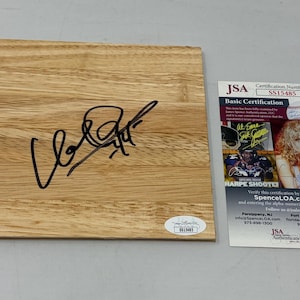 May include: A light-colored wooden plaque with a black signature. A JSA Basic Certification card is next to the plaque. The card features a photo of a woman and text. A small sticker is on the plaque.