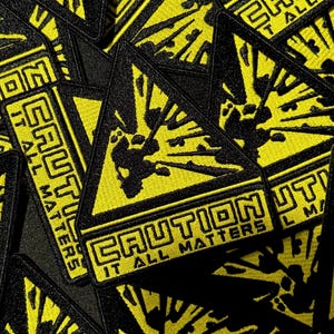 May include: A collection of triangular embroidered patches featuring a yellow background with a black graphic of an explosion. Each patch includes the text CAUTION and IT ALL MATTERS in bold black lettering, with a thick black border surrounding the design.